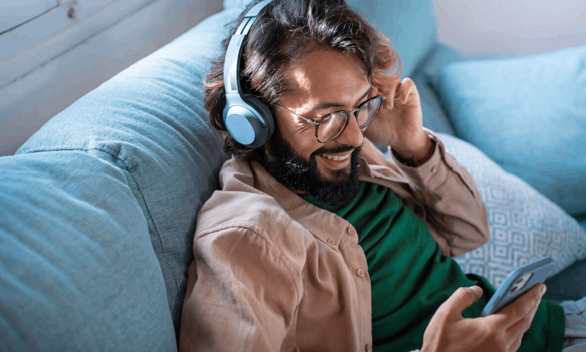 A man wearing headphones and smiling while listening to music on his phone as he relaxes on a couch.